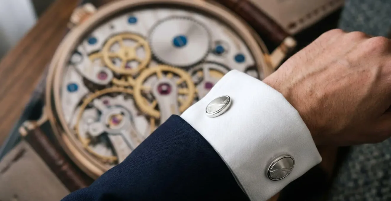 Gros plan artistique de boutons de manchette en argent massif sur une chemise blanche à poignet mousquetaire, avec une montre suisse en arrière-plan flou